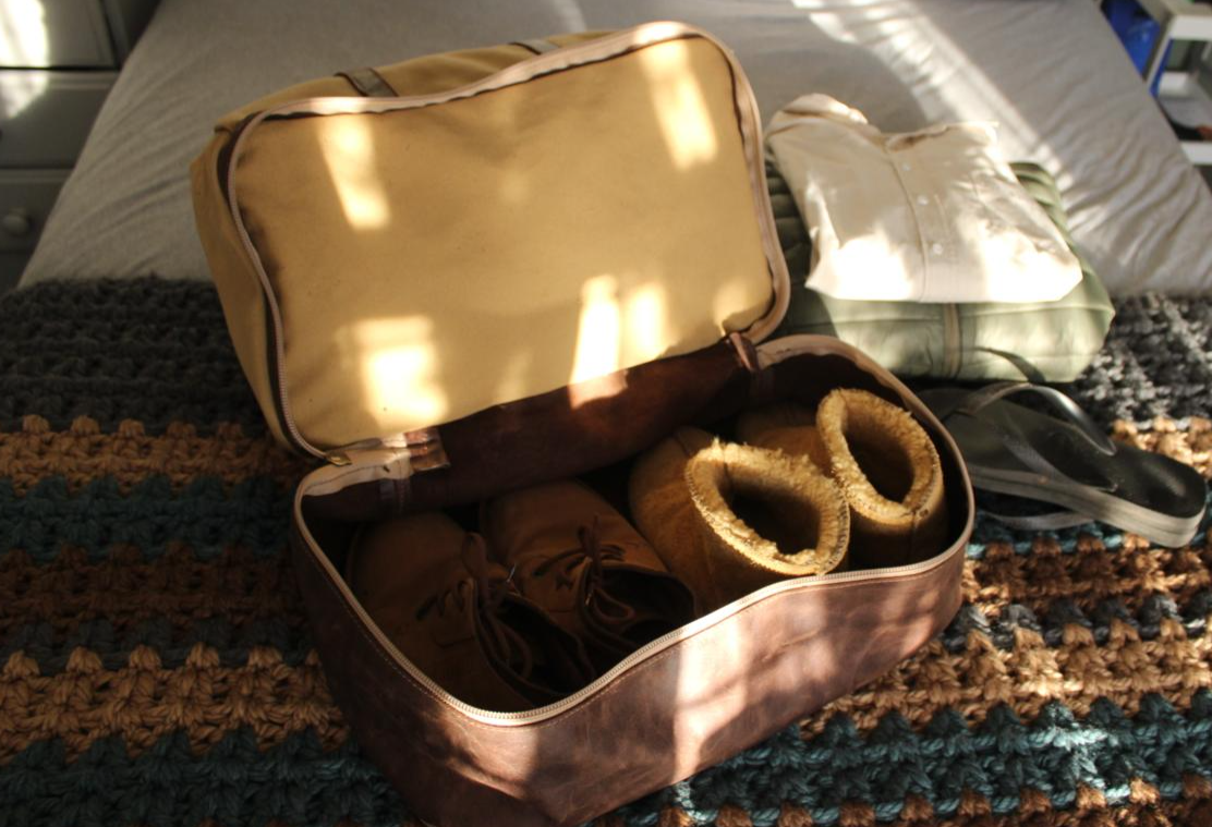 A beige and brown leather travel bag with a shoe pocket, placed on a bed next to a pair of brown boots and a stack of folded green shirts.