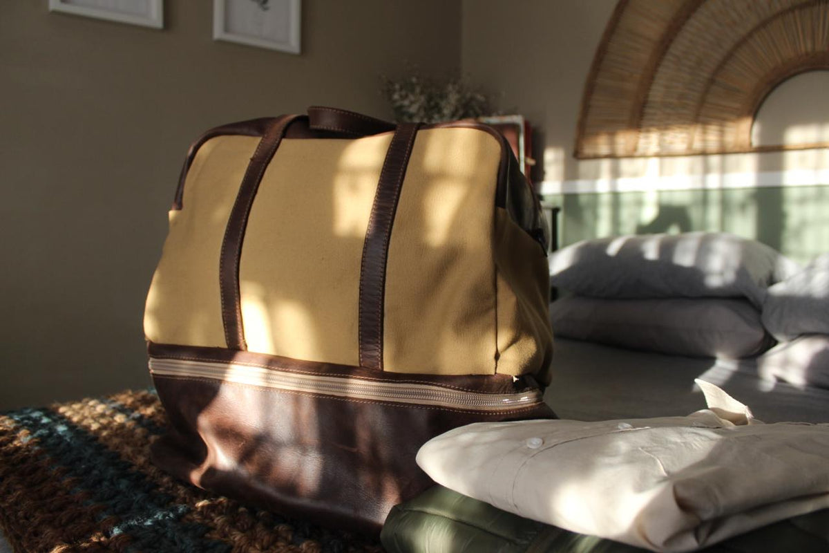 A beige and brown leather travel bag with a shoe pocket, placed on a bed next to a pair of brown boots and a stack of folded green shirts.