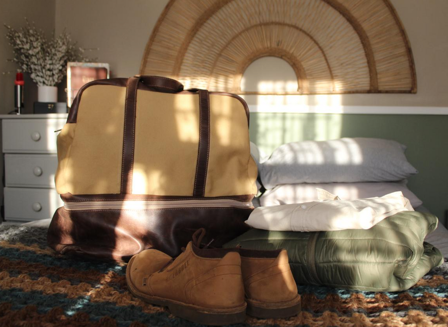 A beige and brown leather travel bag with a shoe pocket, placed on a bed next to a pair of brown boots and a stack of folded green shirts.