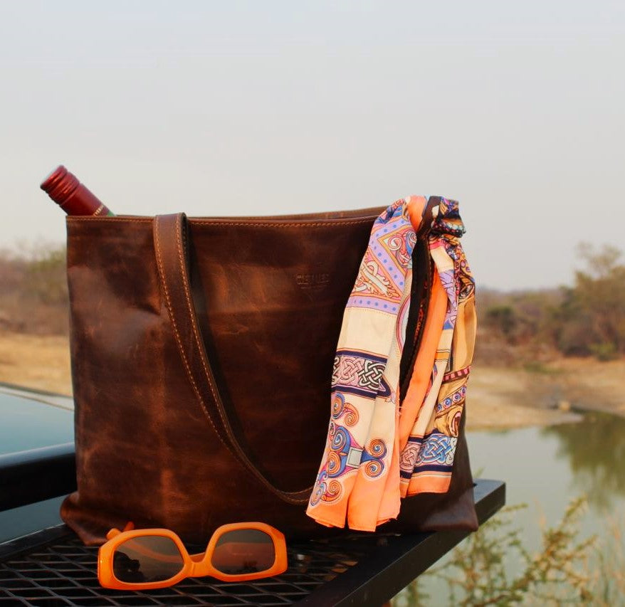 A brown leather tote bag with a multicolored scarf and orange sunglasses placed on a surface, possibly wood, with a natural landscape in the background.