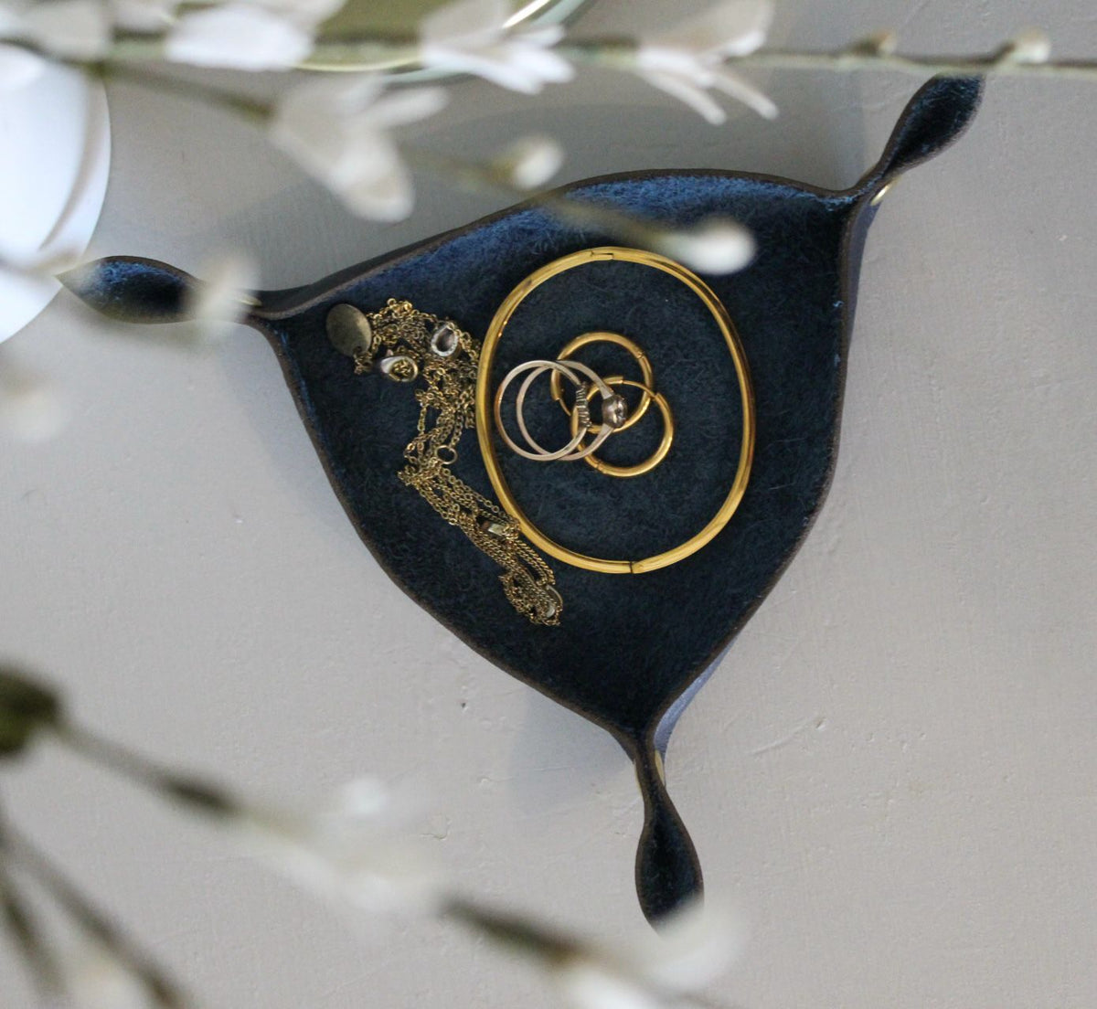 A black leather jewellery holder with a gold-tone chain and ring design, displayed on a white background with floral accents.