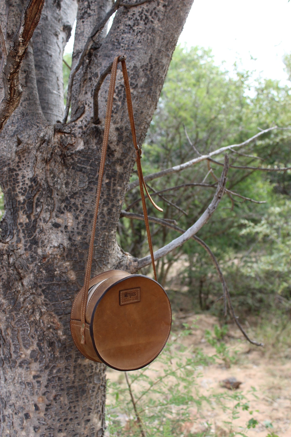 Round, brown leather shoulder bag hanging from a tree branch
