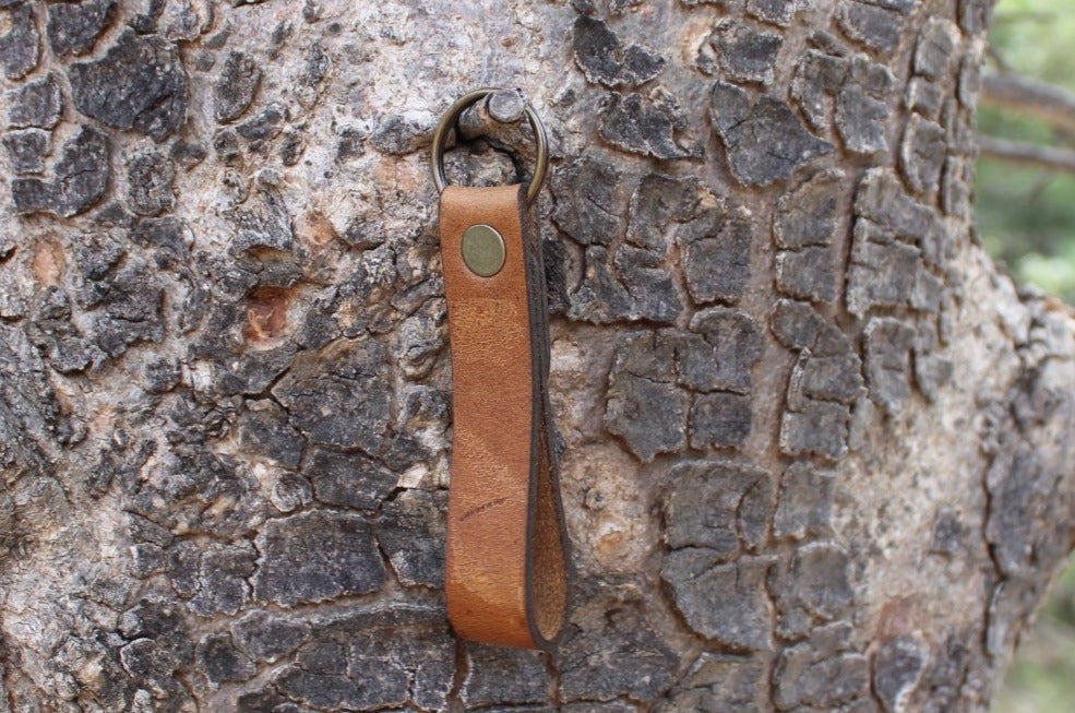 A brown leather keychain with a metal ring and a stamped tree logo, attached to a textured tree bark.