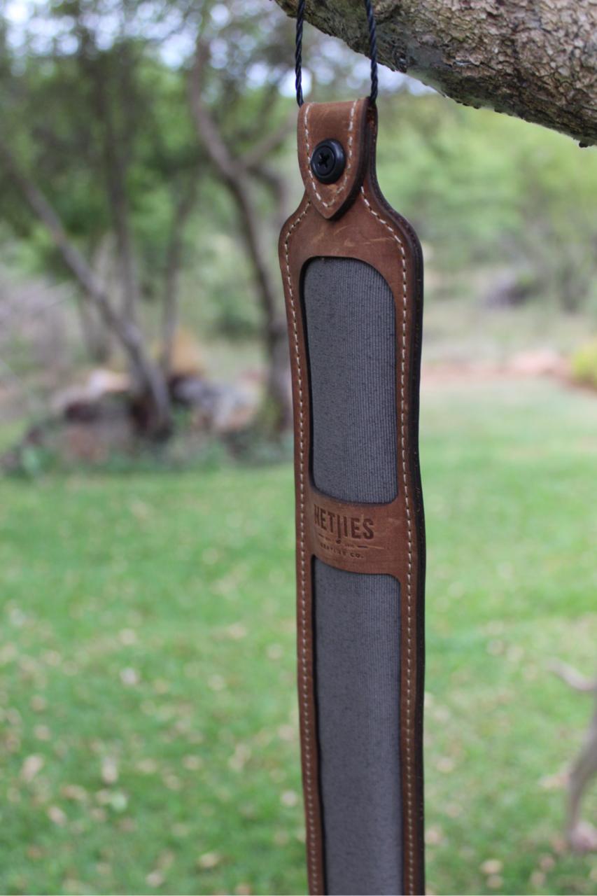 A brown leather rifle sling hanging from a tree branch, with a logo visible on the strap.