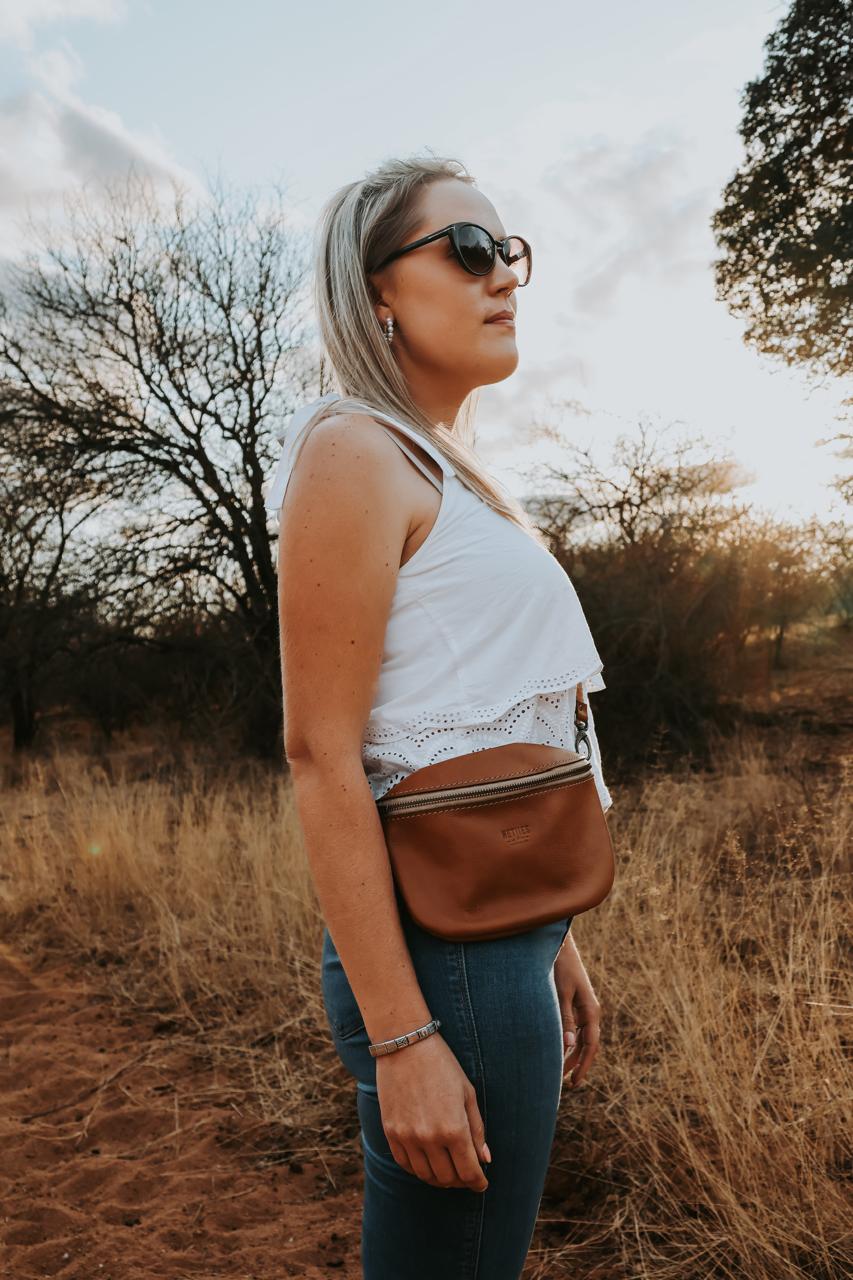 Woman standing in a field wearing sunglasses and a white top with a brown bag.