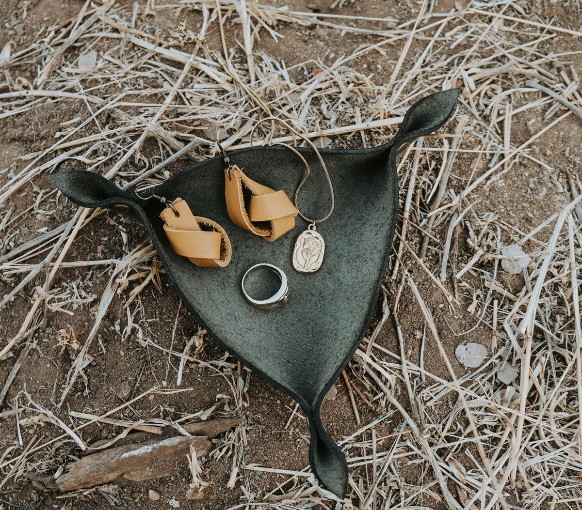 Jewelry including earrings and a ring in a black leather jewelry holder placed on the ground outdoors with natural elements.