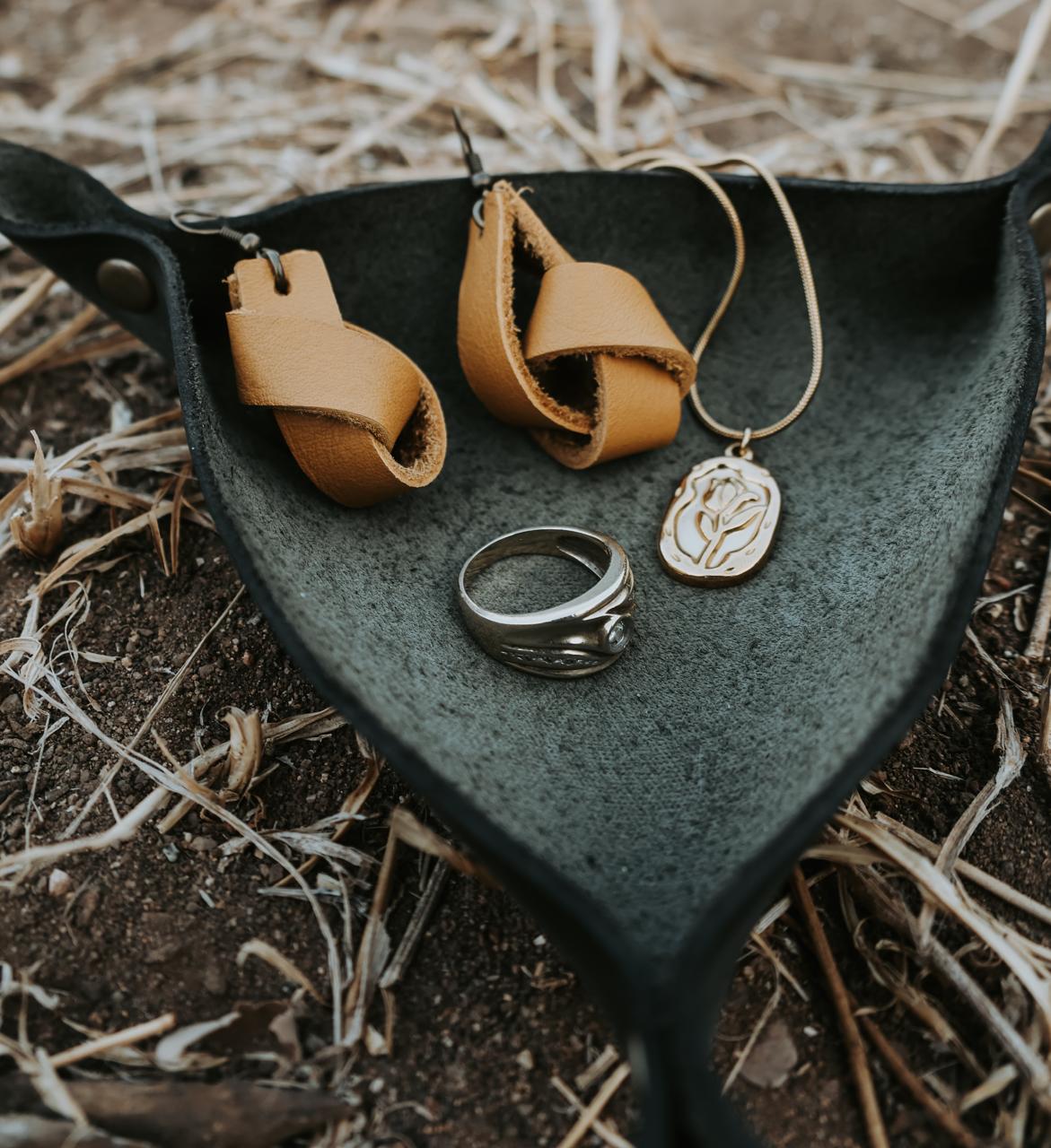 Jewelry including earrings, a ring, and a pendant in a black leather jewelry holder placed outside on the ground with a natural background.