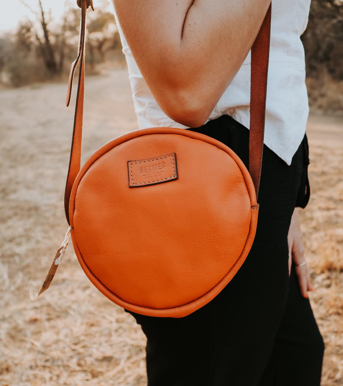 Person holding a round orange leather bag with a visible brand label in an outdoor setting.