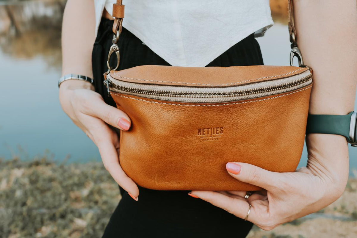Person holding a brown leather bag with 'Netjies' branding by a body of water.