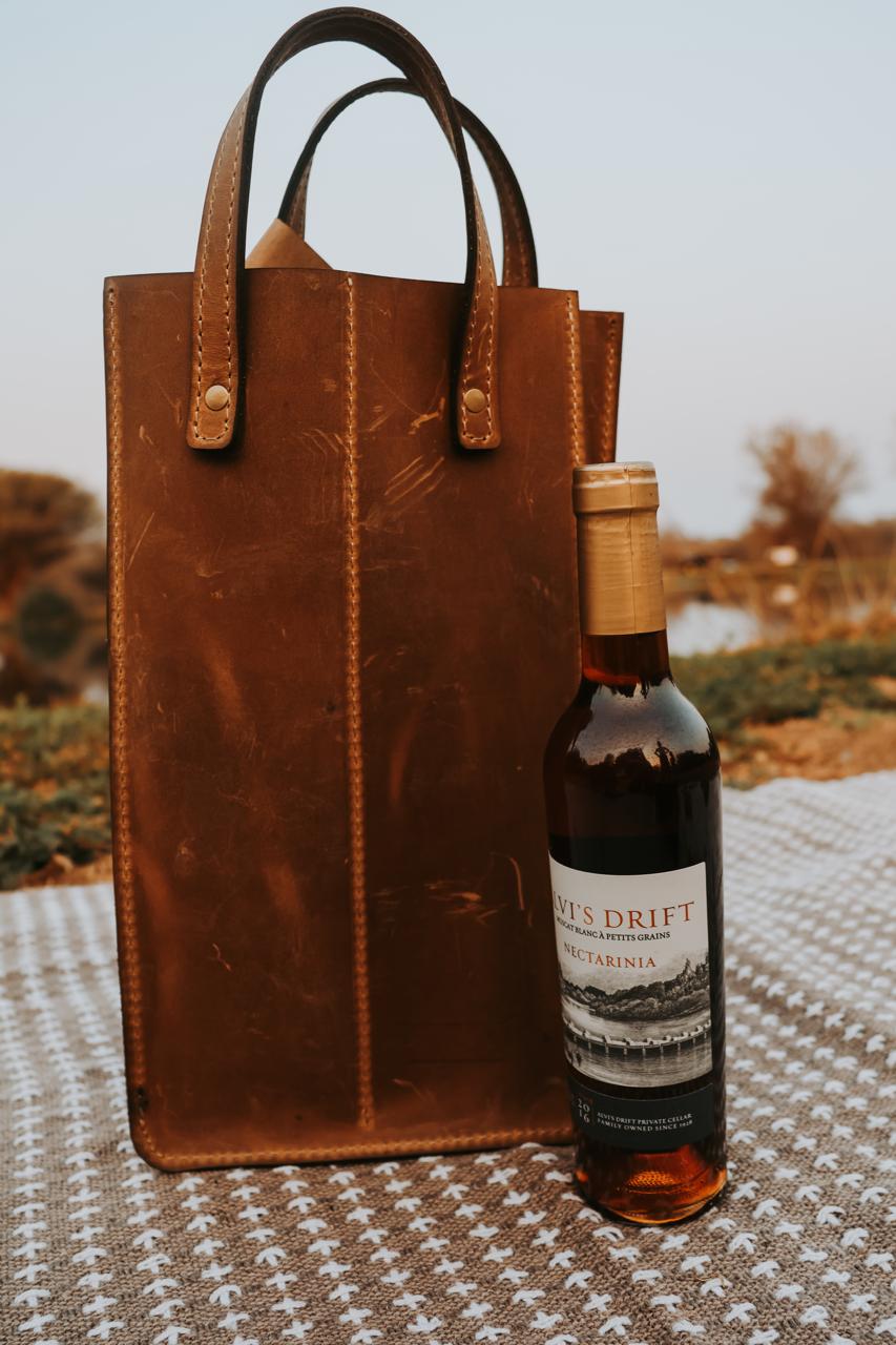 Brown leather wine tote bag with a bottle of wine on a textured surface outdoors.