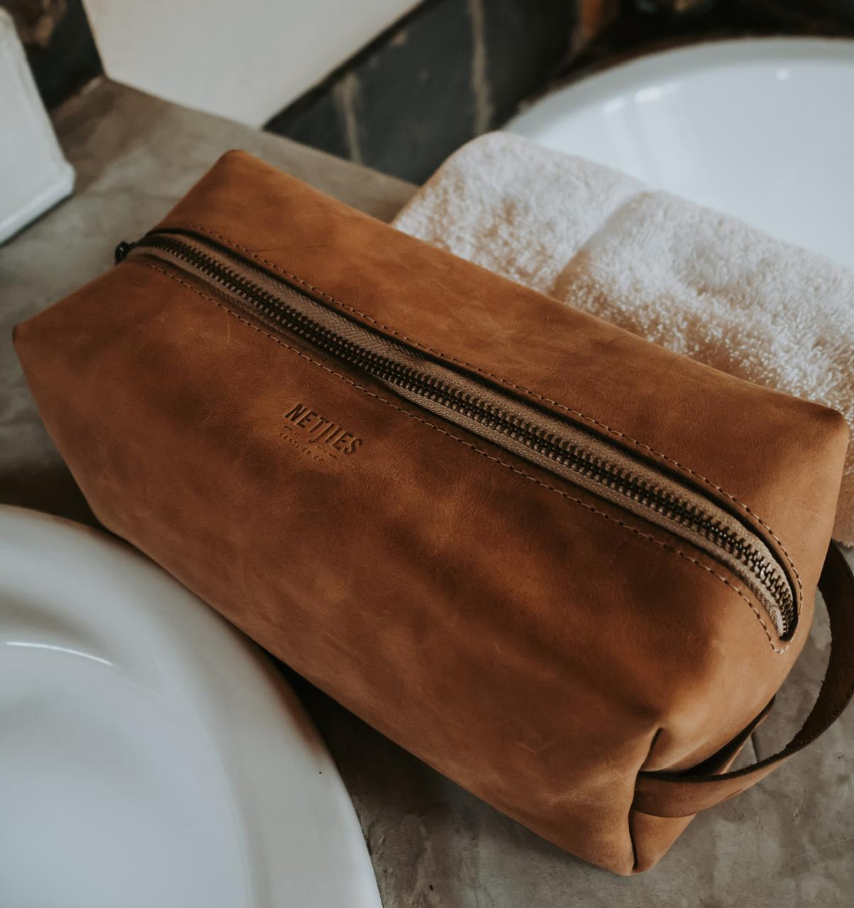 Brown leather toiletry bag with a zipper on a bathroom counter.
